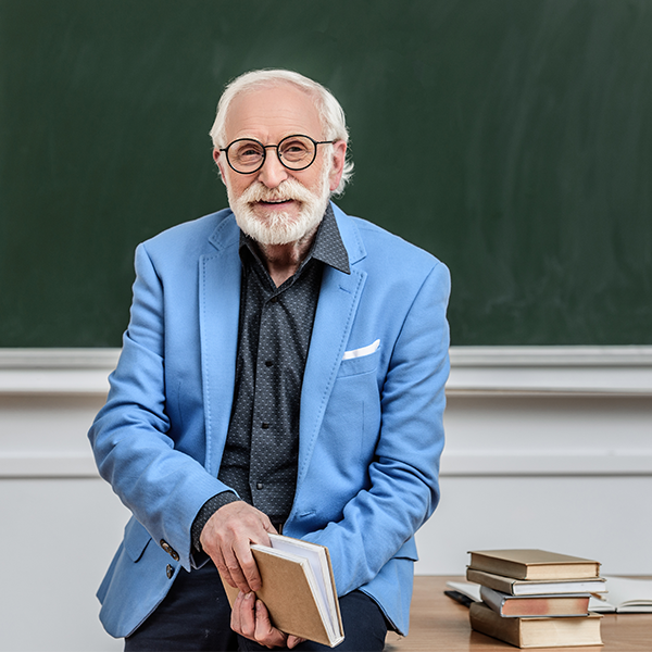 bearded professor in blue jacket in front of a blackboard a book in hand